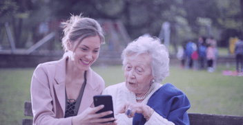 A younger and older person sit together on a park bench, engaging with a smartphone—symbolising intergenerational connection and the role of technology in preserving community values.