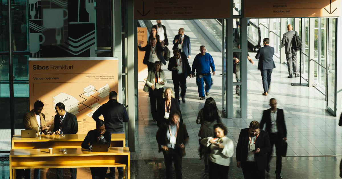 Busy indoor scene at Sibos Frankfurt, showing attendees walking and talking near a large venue overview sign with a map. People are engaged in conversations or using phones, with glass doors and more attendees visible in the background.