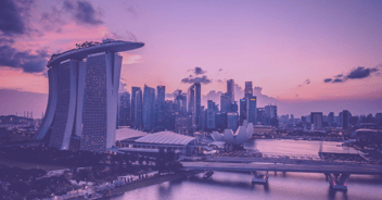 A panoramic view of Singapore's Marina Bay Sands and the city skyline at dusk. The iconic Marina Bay Sands hotel with its boat-shaped rooftop is prominently featured on the left, while the ArtScience Museum, resembling a lotus flower, is visible in front of the skyscrapers. 
