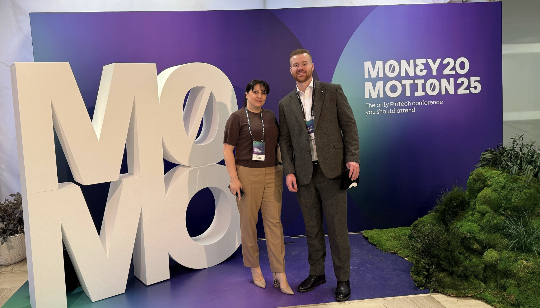  10x Commercial Director William Hatfield stands with another attendee in front of a large display at Money Motion 2025. The backdrop features a purple-blue gradient with the event name and tagline, "The only FinTech conference you should attend"