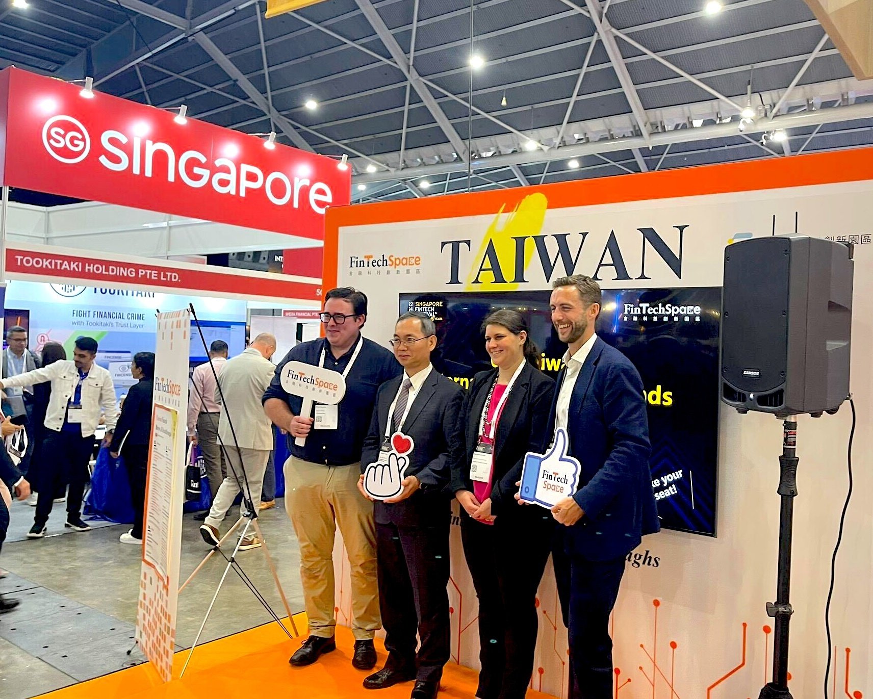 SVP APAC for 10x, Lewis Ide, stands with 3 people in front of a booth at the Singapore FinTech Festival. The booth has an orange frame and a large sign reading “TAIWAN” and “FinTechSpace” at a trade show. They are holding props shaped like social media icons and signs with the FinTechSpace logo. Behind them is a screen displaying text, and to the left is a red overhead banner with “Singapore” in bold white letters. Other exhibition booths and attendees are visible in the background under a high, industrial-style ceiling.